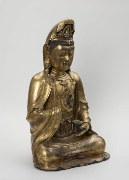 Bodhisattwa Guanyin - ujęcie przodu ze skosu z lewej; Duża, wykonana ze złoconego brązu figura przedstawia siedzącą w pozie medytacyjnej kobietę Bodhisattwę Guanyin (chin. 观音, sanskr. Awalokiteśwara, tyb. Czenrezig). Postać ma spuszczone, przymknięte oczy, symetryczną, pełną twarz o pogodnym i spokojnym wyrazie oraz gładko zaczesane włosy, schowane pod diademem i zaciągniętą na głowę tkaniną. Ubrana jest w długie, warstwowe, jedwabne szaty królewskie oraz wyszukaną, ciężką biżuterię książęcą: diadem, w którego centrum znajduje się niebiański Budda Amitabha (sanskr. Amitābha); pary dużych, okrągłych kolczyków ze spiczastymi zawieszkami oraz grubego naszyjnika z klejnotem, od którego odchodzi siedem pasów korali z wisiorkami. Jej prawa dłoń uniesiona na wysokości klatki piersiowej ułożona jest pionowo, jej środkowy palec i kciuk tworzą okrąg trzymając niezidentyfikowany, haczykowaty przedmiot. Poniżej znajduje się ustawiona poziomo lewa dłoń, która ściska niewielką wazę.