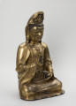 Bodhisattwa Guanyin - ujęcie przodu ze skosu z lewej; Duża, wykonana ze złoconego brązu figura przedstawia siedzącą w pozie medytacyjnej kobietę Bodhisattwę Guanyin (chin. 观音, sanskr. Awalokiteśwara, tyb. Czenrezig). Postać ma spuszczone, przymknięte oczy, symetryczną, pełną twarz o pogodnym i spokojnym wyrazie oraz gładko zaczesane włosy, schowane pod diademem i zaciągniętą na głowę tkaniną. Ubrana jest w długie, warstwowe, jedwabne szaty królewskie oraz wyszukaną, ciężką biżuterię książęcą: diadem, w którego centrum znajduje się niebiański Budda Amitabha (sanskr. Amitābha); pary dużych, okrągłych kolczyków ze spiczastymi zawieszkami oraz grubego naszyjnika z klejnotem, od którego odchodzi siedem pasów korali z wisiorkami. Jej prawa dłoń uniesiona na wysokości klatki piersiowej ułożona jest pionowo, jej środkowy palec i kciuk tworzą okrąg trzymając niezidentyfikowany, haczykowaty przedmiot. Poniżej znajduje się ustawiona poziomo lewa dłoń, która ściska niewielką wazę.