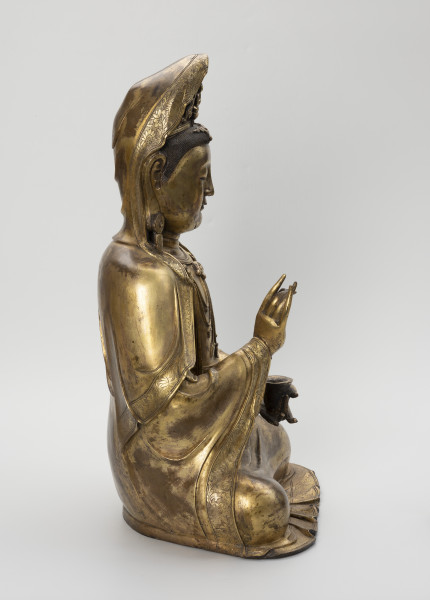 Bodhisattwa Guanyin - ujęcie z boku; Duża, wykonana ze złoconego brązu figura przedstawia siedzącą w pozie medytacyjnej kobietę Bodhisattwę Guanyin (chin. 观音, sanskr. Awalokiteśwara, tyb. Czenrezig). Postać ma spuszczone, przymknięte oczy, symetryczną, pełną twarz o pogodnym i spokojnym wyrazie oraz gładko zaczesane włosy, schowane pod diademem i zaciągniętą na głowę tkaniną. Ubrana jest w długie, warstwowe, jedwabne szaty królewskie pary dużych, okrągłych kolczyków ze spiczastymi zawieszkami. Jej prawa dłoń uniesiona na wysokości klatki piersiowej ułożona jest pionowo. Poniżej znajduje się ustawiona poziomo lewa dłoń, która ściska niewielką wazę.