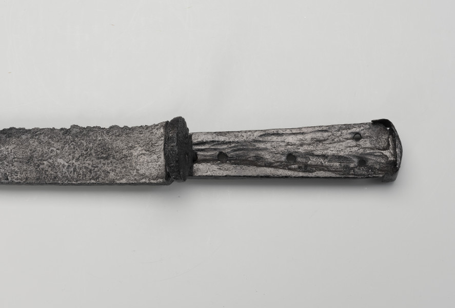 kord - Ujęcie 3/4 z góry; Głownia kordu jest prosta, jednosieczna, o sztychu decentrycznym. Trzon z jednostronnym wyżłobionym wgłębieniem ma cztery otwory do mocowania okładzin rękojeści, których brak. Dwa ostatnie otwory w trzonie umiejscowione tuż przy kapturku głowicy wydrążone są w pionowym ustawieniu. Trzon oddzielony jest od głowni nasadą, przy niej zamocowana jest tarczka w formie muszli. Głowica jest w formie kapturka, czyli cienkiej stalowej blaszki wygiętej w kierunku ostrza.