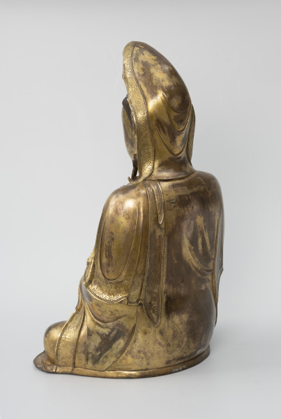 Bodhisattwa Guanyin - ujęcie tyłu ze skosu z lewej; Duża, wykonana ze złoconego brązu figura przedstawia siedzącą w pozie medytacyjnej kobietę która ma zaciągniętą na głowę tkaniną. Ubrana jest w długie, warstwowe, jedwabne szaty królewskie.
