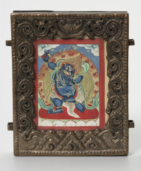 relikwiarz gau z miniaturą sakralną tsakli - ujęcie z przodu; Miedziany relikwiarz w formie małej, wąskiej, przeszklonej i bogato zdobionej ramki wypełnionej obrazem Wadżrapani (sanskr. Vajrapāṇi, tyb. Cziano Dordże). Postać zaprezentowana w  formie gniewnej, najprawdopodobniej przedstawia Wadżrapani jako strażnika Dharmy (sanskr. dharamapāla) – Wadżrapani-Acharya. Ukazany jako tęgi i silny, z potężnym ciemnoniebieskim ciałem, wystającym brzuchem, grubymi, krótkimi kończynami i wściekłym wyrazem twarzy. W odmianie ikonograficznej z jedną głową, trzecim okiem, nastroszonymi włosami, pięcioramienną koroną bodhisattwy, naszyjnikami z węży oraz pasem z tygrysiej skóry. Otoczony płomieniami trzyma miecz i lasso (sanskr. pāśa). Stoi na lotosie – symbolu czystości i poznania, na tle nieba i chmur. Obrazek zakończony czerwoną bordiurą. Ramka ozdobiona prostymi nacięciami przy zewnętrznych krawędziach oraz misternie wykonanymi wzorami przy wewnętrznych: usytuowany po bokach wzór grawerowanej wici roślinnej, centralnie, w górnej części znajduje się przedstawienie przypominające baldachim (parasol buddyjski), natomiast zlokalizowane w dolnej części trójkątne motywy być może przedstawiają muszle - motyw konchy. Przy bocznych powierzchniach dwa prostokątne mocowania - ucha na pasek do noszenia relikwiarza, ułożone symetrycznie. Widoczne ślady użytkowania, liczne przetarcia, odbarwienia.