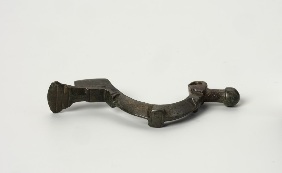 fibula z brązu z Borkowic typu Bügelknopffibel - Ujęcie z góry; Zapinka z brązu z guzkiem na łukowatym kabłąku typu Bügelknopffibel. Fibula pozbawiona jest sprężynki i szpili. Kabłąk jest w trzech miejscach zdobiony profilowanymi guzkami w symetrycznym układzie.