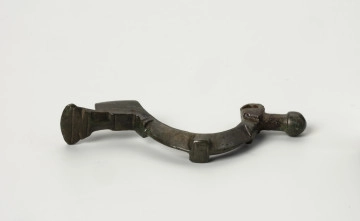 MNS/A/7135/2 fibula z brązu z Borkowic typu Bügelknopffibel - Ujęcie z góry; Zapinka z brązu z guzkiem na łukowatym kabłąku typu Bügelknopffibel. Fibula pozbawiona jest sprężynki i szpili. Kabłąk jest w trzech miejscach zdobiony profilowanymi guzkami w symetrycznym układzie.