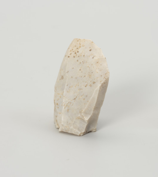 rdzeń stożkowy - Ujęcie z przodu; Rdzeń z bryły krzemienia kredowego bardzo wysokiej jakości w końcowym stadium eksploatacji o znacznym stopniu redukcji uniemożliwiającym jego utrzymanie. Forma zbliżona do stożka lub ostrosłupa o dookolnej odłupni i płaskiej, lekko wklęsłej pięcie uformowanej jednym odbiciem. Widoczny na jednej ze stron duży negatyw wskazuje na naprawę rdzenia krótko przed zakończeniem eksploatacji. W masie widoczne są liczne długie egzoszkielety organizmów morskich, ułożone w podłużne linie. Zabytek w całości pokryty jest białą patyną wskazującą na długotrwałe zaleganie na powierzchni.