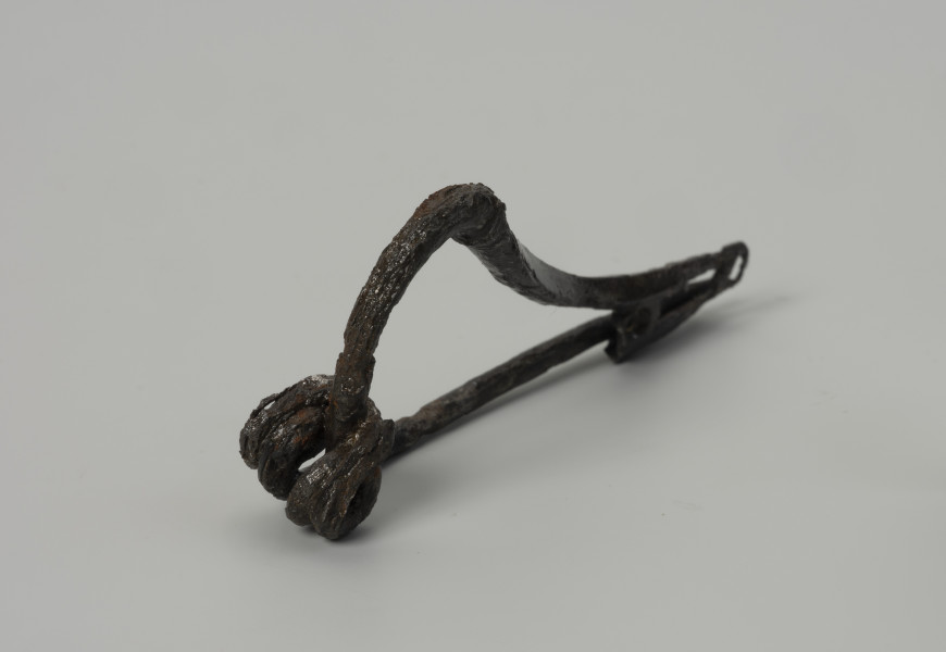 fibula wariant N wg Kostrzewskiego - Ujęcie przodu ze skosu z prawej; Żelazna zapinka wariantu N wg Kostrzewskiego, z wysokim kabłąkiem z okrągłego drutu, z guzem plastycznym. Sprężynka z czterema zwojami i dolną cięciwą. Ażurowa pochewka ma dwa otwory.