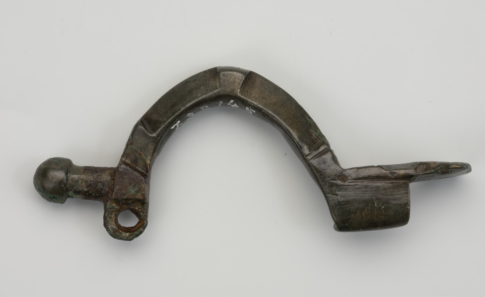 fibula z brązu z Borkowic typu Bügelknopffibel - Ujęcie z boku; Zapinka z brązu z guzkiem na łukowatym kabłąku typu Bügelknopffibel. Fibula pozbawiona jest sprężynki i szpili.