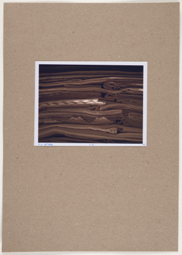 fotografia - Barwna odbitka fotograficzna w stonowanych kolorach przedstawiająca stos poskładanych tkanin leżących jedna na drugiej. Brak widocznych zakończeń tkanin wskazuje, że jest to kadr większej całości. Fotografia utrzymana jest w tonacji brązowo-beżowej z jasną plamą świetlną widoczną w centralnej części. Tkaniny leżą warstwami, płasko, w niektórych miejscach widoczne jest zawinięcie lub miejsca złożenia którejś z nich. W dwóch miejscach (w tym jednym z jaśniejszy oświetleniem), widoczny jest brzeg ozdobiony haftem i mereżką. Odbitka fotograficzna została wykonana z wąską, białą ramką.