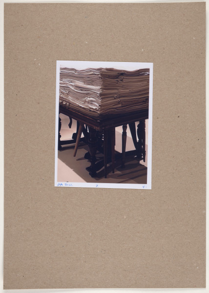fotografia - Barwna odbitka fotograficzna utrzymana w stonowanych kolorach, w tonacji beżowo-brązowej, przedstawiająca w centralnej części ciemny, prostokątny stół o masywnym blacie z ryflowanymi krawędziami stojący na czterech ozdobnych nogach, które są połączone ze sobą parami, za pomocą listew umocowanych tuż nad podłogą. Do stołu zamontowanych zostało nierównomiernie kilka innych nóg pochodzących z innych mebli, o innych kolorach, o różnej długości i kształcie, niektóre z ich są ozdobne, inne - proste. Na blacie stołu umieszczony został duży stos poskładanych tkanin w różnych odcieniach beżu, leżących jedna na drugiej. Cała instalacja stoi we wnętrzu na jasnym tle, na podłodze pod nią widoczny jest rzucany przez nią cień. Odbitka fotograficzna została wykonana z wąską, białą ramką.