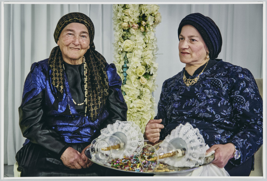 scena rodzajowa - Poziomy, barwny kadr fotograficzny przedstawiający portrety 2 dojrzałych kobiet ubranych w ciemne, ozdobne stroje, czarne chusty zawiązane dookoła głów. Chusta kobiety po lewej stronie ma dekoracyjny, złoty, geometryczny wzór. Kobiety trzymają na kolanach dużą metalową tacę z leżącymi na niej dwoma świecami o szklanych kloszach. Pomiędzy lampionami nasypane jest kolorowe, metalizujące konfetti. Kobiety siedzą na tle białej udrapowanej zasłony i dwóch słupów udekorowanych białymi kwiatami.