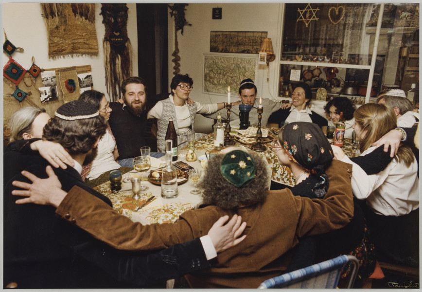 fotografia - Zdjęcie przedstawia grupę osób siedzących przy zastawionym stole podczas wieczerzy. Siedzą na przemian, mężczyźni i kobiety. Mężczyźni mają na głowach jarmułki. Jedna z kobiet (po prawej stronie kadru) ma na głowie ciemną chustę. Wszyscy obejmują się nawzajem ramionami. Nas stole stoją min. dwie zapalone świece w wysokich świecznikach, wino w pucharkach oraz talerz z macą. Ściany mieszkania zdobią makaty, mapy oraz fotografie. Po prawej stronie kompozycji widoczne okno, a na jego szybie symbol sześcioramiennej gwiazdy (gwiazdy Dawida).