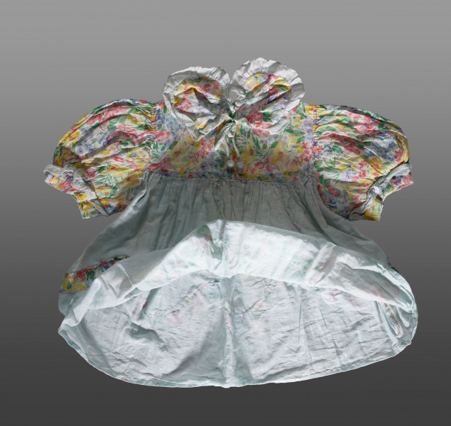 Sukienka dziecięca wykonana z tkaniny bawełnianej w dwóch kolorach. Dolna część sukienki uszyta jest z tkaniny w kolorze jasnobłękitnym. Karczek, rękawki i kołnierzyk wykonane z tkaniny ozdobionej nadrukiem w kolorowe kwiaty. Rękawki (bufiaste) i kołnierzyk wykończone ozdobną lamówką z tkaniny w kolorze jasnobłękitnym. Kołnierzyk wiązany na cienki sznureczek w kolorze lamówki. U dołu sukienki (około 1/3 wysokości od dolnej krawędzi) naszyta ozdobna lamówka z tkaniny w kwiaty. Lamówka układa się w falisty wzór.