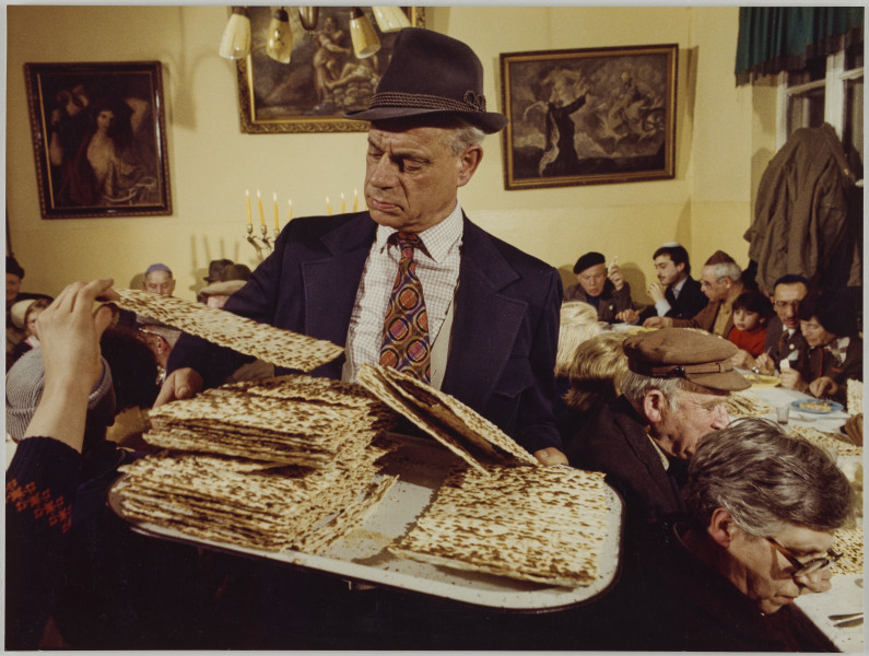 fotografia - Zdjęcie przedstawia grupę osób podczas wieczerzy. Na pierwszym planie (przodem do obiektywu) widoczny mężczyzna w garniturze, białej koszuli w drobna kratę, wzorzystym krawacie oraz kapeluszu. Trzyma przed sobą tacę, na której ułożona jest maca. Po lewej stronie kadru widać wyciągniętą rękę, która zabiera z tacy jeden kawałek macy. W tle widać stały ustawione w podkowę i nakryte białym obrusem. Stoły są zastawione pożywieniem a przy nich (po obu stronach) siedzą biesiadnicy. Za mężczyzną (na środkowym stole) widoczny fragment wieloramiennego świecznika, płonące w nim świece oraz żółtą ścianę, na której zawieszone są trzy obrazy. W górze kadru widoczny fragment żyrandola. 