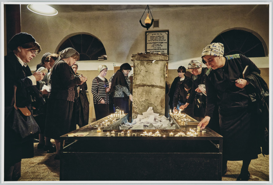 scena rodzajowa - Poziomy, barwny kadr fotograficzny przedstawiający grupę kobiet zebranych wokół nagrobka znajdującego się pośrodku pomieszczenia o dwóch półokrągłych oknach. Jest ono oświetlone dwoma lampami. Kobiety są ubrane w podobne stroje w stonowanych kolorach (spódnica za kolano, bluzka, żakiet lub sweter) oraz chusty lub czapki na głowach. Część z nich zapala niewielkie świece (tzw. tealighty) na stolikach otaczających nagrobek, część stoi ze spuszczona głową w pozie sugerującej skupienie/modlitwę.