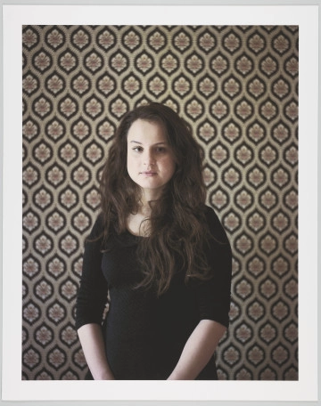 portret fotograficzny - Fotografia barwna skadrowana pionowo, przedstawiająca półpostaciowy portret młodej kobiety. Portretowana dziewczyna o ciemnych, długich, falujących włosach, puszczonych luźno na ramiona, ubrana w ciemną prostą bluzkę z długimi rękawami podciągniętymi pod łokcie, patrzy prosto w stronę widza. Kobieta stoi we wnętrzu na tle ściany pokrytej tapetą o bardzo ozdobnym, symetrycznym, floralno-geometrycznym wzorze.
