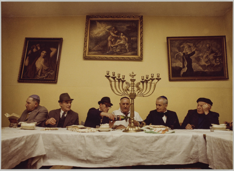 fotografia - Zdjęcie przedstawia grupę sześciu mężczyzn siedzących za stołem podczas wieczerzy sederowej. Mężczyźni siedzą po jednej stronie stołu, pod ścianą. Po środku stołu widoczny ozdobny ośmioramienny świecznik (miejsca na świece są puste). Stół jest nakryty białym obrusem i zastawiony potrawami. Mężczyźni są ubrani w garnitury oraz nakrycia głowy (jarmułki lub kapelusze). Trzeci mężczyzna od lewej nalewa wino do kieliszka drugiego mężczyzny od prawej strony. W tle, na ścianie widoczne trzy obrazy w ozdobnych ramach.