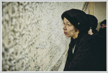 scena rodzajowa - Poziomy, barwny kadr fotograficzny przedstawiający popiersiowy portret fotograficzny dojrzałej kobiety w ubranej w czarną bluzkę, czarną czapkę i srebrny naszyjnik stojącej przed białą, koronkową zasłoną i patrzącą z zamyśleniem przed siebie.