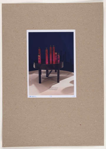 fotografia - Barwna odbitka fotograficzna przedstawiająca ciemny stół o masywnym blacie stojący na trzech walcowatych nogach. Do stołu zamontowane zostały kolejne dwie nogi, które są krótsze, zatem nie stanowią podpór. Na blatu stołu widocznych jest siedem wysokich, cylindrycznych elementów o różnej wysokości, ustawionych nieregularnie. Są to walce zakończone połyskliwymi, ciemnymi nasadkami, z przeźroczystego materiału o częściowo transparentnym wypełnieniu w nasyconych barwach (czerwono-pomarańczowych-różowych). W środku widoczne są pęcherze powietrza. Cała instalacja stoi we wnętrzu na tle ciemnej ściany i na białej podłodze, na której widoczny jest kilkuwersowy czarny napis umieszczony przed obiektem. Odbitka fotograficzna została wykonana z wąską, białą ramką.