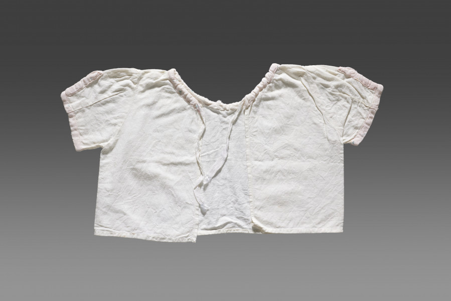 Kaftanik dziecięcy wykonany z cienkiej tkaniny bawełnianej w kolorze białym/écru. Kaftanik o prostym kroju i krótkim rękawku. Rękawy oraz linia dekoltu (brak kołnierzyka) wykończone jasnoróżową tasiemką z widocznym nadrukiem floralnym. Kaftanik wiązany pod szyją na białą tasiemkę. 