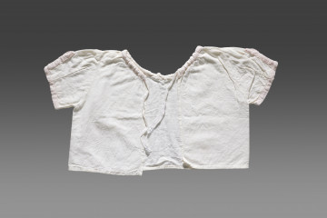 Kaftanik dziecięcy wykonany z cienkiej tkaniny bawełnianej w kolorze białym/écru. Kaftanik o prostym kroju i krótkim rękawku. Rękawy oraz linia dekoltu (brak kołnierzyka) wykończone jasnoróżową tasiemką z widocznym nadrukiem floralnym. Kaftanik wiązany pod szyją na białą tasiemkę. 