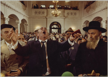 MPOLIN-M1581 fotografia - Zdjęcie przedstawia wnętrze synagogi oraz modlących się w niej mężczyzn, którzy siedzą w kolejnych rzędach ławek. Po środku kompozycji, w pierwszej ławce widoczny mężczyzna z rozłożonymi szeroko rękami i otwartymi ustami. Ma na sobie garnitur, białą koszulę i granatowy krawat w drobne kropki. Na ławce obok niego siedzi dwóch mężczyzn (po lewej i prawej stronie). Mężczyzna po prawej ma na sobie kapelusz z szerokim rondem oraz garnitur i białą koszulę. Ma długą siwą brodę. Mężczyzna po lewej stronie ma na sobie białą koszulę, jasny krawat oraz beżowy płaszcz. Na głowie ma czarny beret. Za nimi widać rzędy ławek, w których siedzą pozostali mężczyźni.