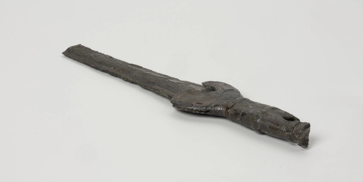 fragment miecza z brązu - Ujęcie z tyłu; Miecz z brązu zachowany we fragmencie - część głowni oraz rękojeść - o łącznej długości 22,7 cm. Egzemplarz cechuje smukła głownia, na powierzchni której przetrwała miejscami pierwotna lśniąca powłoka barwy ciemnozielonej. Dzięki temu widoczne są po obu stronach ostrza struziny podkreślone delikatnym zdobieniem w postaci drobnych nacięć i łuczków. Podobnym ornamentem, na który składają się pasma linii rytych i niewielkich nacięć, pokryta jest również rękojeść miecza o gładkiej ciemnozielonej powierzchni. Rękojeść jest niekompletna, brakuje jej głowicy – najpewniej tworzyły ją dwie niewielkie woluty, pomiędzy którymi wystawał niewielki pręt.