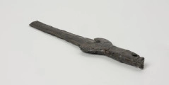 fragment miecza z brązu - Ujęcie z tyłu; Miecz z brązu zachowany we fragmencie - część głowni oraz rękojeść - o łącznej długości 22,7 cm. Egzemplarz cechuje smukła głownia, na powierzchni której przetrwała miejscami pierwotna lśniąca powłoka barwy ciemnozielonej. Dzięki temu widoczne są po obu stronach ostrza struziny podkreślone delikatnym zdobieniem w postaci drobnych nacięć i łuczków. Podobnym ornamentem, na który składają się pasma linii rytych i niewielkich nacięć, pokryta jest również rękojeść miecza o gładkiej ciemnozielonej powierzchni. Rękojeść jest niekompletna, brakuje jej głowicy – najpewniej tworzyły ją dwie niewielkie woluty, pomiędzy którymi wystawał niewielki pręt.