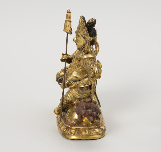 Wajśrawana (Dzambala) - ujęcie z prawej; Złota, męska postać siedząca bokiem, z lewą nogą opuszczoną w dół (poza lalitasana), na złotym, ryczącym lwie. W prawej ręce trzyma chorągiew zwycięstwa (osobny, wyciągany element), a w lewej zwierzątko: mangustę  (ichneumon), która wypluwa klejnot z pyszczka. Postać w gniewnej formie ma upięte w kok czarne włosy, szlachetnie ubrana: nosi diadem, liczne ozdoby z klejnotów, a jej strój przypomina zbroję wysokiej rangi dostojnika/króla. Lwa o bordowych brwiach, sierści na głowie i ogonie przedstawiono w pozycji wpółleżącej. Obie postacie ustawiono na lotosowej podstawie. W jej centralnym punkcie wydrążono niewielki otwór na szpic sztandaru. Posążek najprawdopodobniej przedstawia bóstwo bogactwa Wajśrawanę wraz z jego atrybutami.