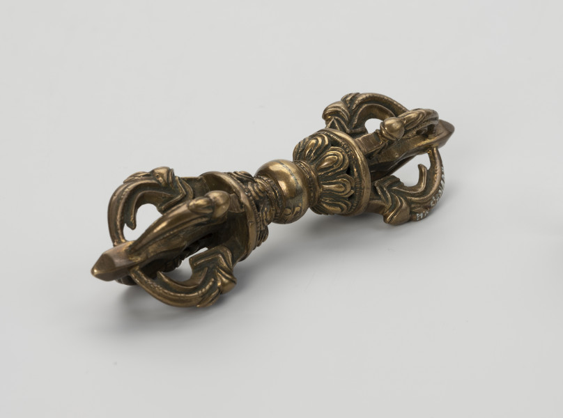 wadżra - ujęcie przodu ze skosu z lewej; Mały, wykonany z brązu, przypominający berło przedmiot w kształcie kokardy lub ósemki. W jego centrum znajduje się kula - uchwyt berła. Po jej obu stronach znajdują się wyrastające w przeciwnych kierunkach stożkowe zgrubienia stylizowane na kwiaty lotosu z ośmioma płatkami. Od każdego z nich odchodzą cztery, łukowate ramiona zbiegające się wokół centralnej szpicy przebiegającej przez całą oś obiektu. Ramiona bogato zdobione w formie ozdoby przypominającej podwinięty liść przedstawiającej wystylizowaną głową potwora zwanego makarą.