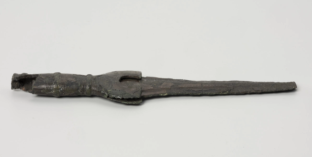 fragment miecza z brązu - Ujęcie z boku; Miecz z brązu zachowany we fragmencie - część głowni oraz rękojeść - o łącznej długości 22,7 cm. Egzemplarz cechuje smukła głownia, na powierzchni której przetrwała miejscami pierwotna lśniąca powłoka barwy ciemnozielonej. Dzięki temu widoczne są po obu stronach ostrza struziny podkreślone delikatnym zdobieniem w postaci drobnych nacięć i łuczków. Podobnym ornamentem, na który składają się pasma linii rytych i niewielkich nacięć, pokryta jest również rękojeść miecza o gładkiej ciemnozielonej powierzchni. Rękojeść jest niekompletna, brakuje jej głowicy – najpewniej tworzyły ją dwie niewielkie woluty, pomiędzy którymi wystawał niewielki pręt.