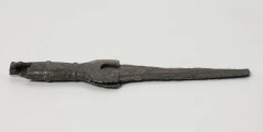 fragment miecza z brązu - Ujęcie z boku; Miecz z brązu zachowany we fragmencie - część głowni oraz rękojeść - o łącznej długości 22,7 cm. Egzemplarz cechuje smukła głownia, na powierzchni której przetrwała miejscami pierwotna lśniąca powłoka barwy ciemnozielonej. Dzięki temu widoczne są po obu stronach ostrza struziny podkreślone delikatnym zdobieniem w postaci drobnych nacięć i łuczków. Podobnym ornamentem, na który składają się pasma linii rytych i niewielkich nacięć, pokryta jest również rękojeść miecza o gładkiej ciemnozielonej powierzchni. Rękojeść jest niekompletna, brakuje jej głowicy – najpewniej tworzyły ją dwie niewielkie woluty, pomiędzy którymi wystawał niewielki pręt.