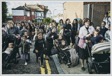 scena rodzajowa - Poziomy, barwny kadr fotograficzny przedstawiający dużą grupę, w której skład wchodzą kobiet, i dziewczęta. Trzy spośród nich, na pierwszym planie, pchają wózki z małymi dziećmi. Grupa zajmuje całą szerokość chodnika i ulicy pomiędzy niewysokimi miejskimi budynkami - idą w kierunku widza.