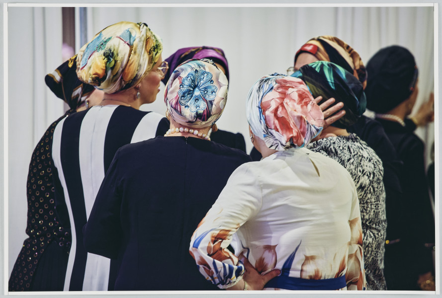 scena rodzajowa - Poziomy, barwny kadr fotograficzny przedstawiający grupę 7 kobiet stojących we wnętrzu, w kręgu i rozmawiających. Kobiety ubrane są w odświętne sukienki oraz kolorowe, wzorzyste chusty zawiązane dookoła ich głów. Ich twarze są niewidoczne dla widza.