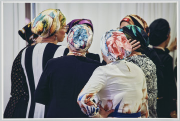 scena rodzajowa - Poziomy, barwny kadr fotograficzny przedstawiający grupę 7 kobiet stojących we wnętrzu, w kręgu i rozmawiających. Kobiety ubrane są w odświętne sukienki oraz kolorowe, wzorzyste chusty zawiązane dookoła ich głów. Ich twarze są niewidoczne dla widza.