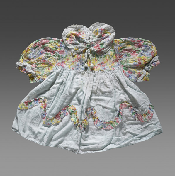 Sukienka dziecięca wykonana z tkaniny bawełnianej w dwóch kolorach. Dolna część sukienki uszyta jest z tkaniny w kolorze jasnobłękitnym. Karczek, rękawki i kołnierzyk wykonane z tkaniny ozdobionej nadrukiem w kolorowe kwiaty. Rękawki (bufiaste) i kołnierzyk wykończone ozdobną lamówką z tkaniny w kolorze jasnobłękitnym. Kołnierzyk wiązany na cienki sznureczek w kolorze lamówki. U dołu sukienki (około 1/3 wysokości od dolnej krawędzi) naszyta ozdobna lamówka z tkaniny w kwiaty. Lamówka układa się w falisty wzór.