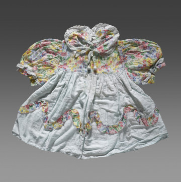 Sukienka dziecięca wykonana z tkaniny bawełnianej w dwóch kolorach. Dolna część sukienki uszyta jest z tkaniny w kolorze jasnobłękitnym. Karczek, rękawki i kołnierzyk wykonane z tkaniny ozdobionej nadrukiem w kolorowe kwiaty. Rękawki (bufiaste) i kołnierzyk wykończone ozdobną lamówką z tkaniny w kolorze jasnobłękitnym. Kołnierzyk wiązany na cienki sznureczek w kolorze lamówki. U dołu sukienki (około 1/3 wysokości od dolnej krawędzi) naszyta ozdobna lamówka z tkaniny w kwiaty. Lamówka układa się w falisty wzór.