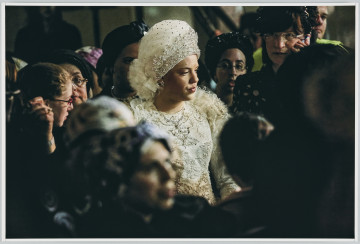 scena rodzajowa - Poziomy, barwny kadr fotograficzny przedstawiający popiersiowy portret panny młodej w centralnej części kadru. Kobieta ubrana jest dekorowaną aplikacjami białą suknię z długimi rękawami i pod szyję oraz bardzo dekoracyjny biały czepiec dekorowany koronką i wyszywany dżetami. Kobieta stoi wśród grupy innych, ubranych w ciemne stroje kobiet, których sylwetki są częściowo nie ostre, szczególnie te na pierwszym – zbliżenie i ostrość ustawiona została na sylwetkę panny młodej.