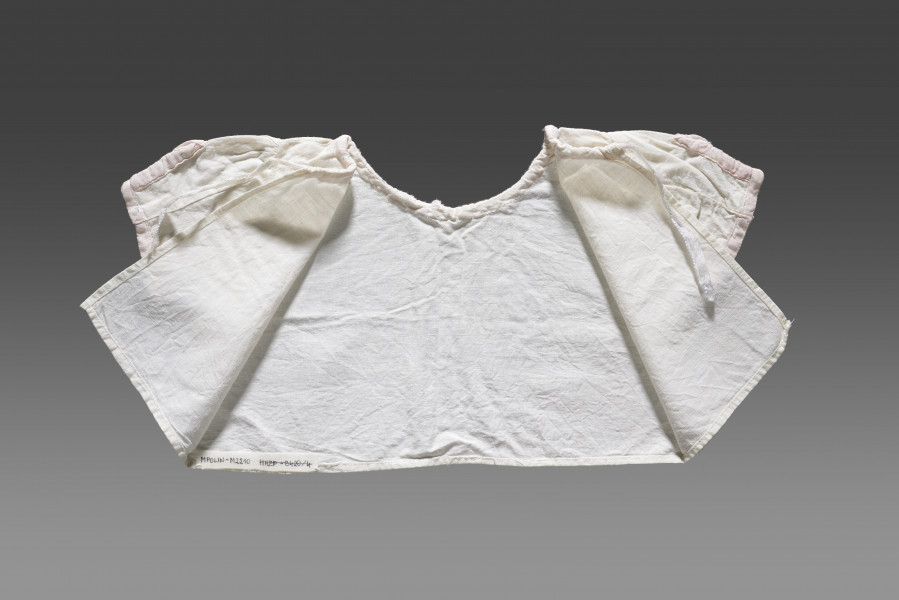 Kaftanik dziecięcy wykonany z cienkiej tkaniny bawełnianej w kolorze białym/écru. Kaftanik o prostym kroju i krótkim rękawku. Rękawy oraz linia dekoltu (brak kołnierzyka) wykończone jasnoróżową tasiemką z widocznym nadrukiem floralnym. Kaftanik wiązany pod szyją na białą tasiemkę. 