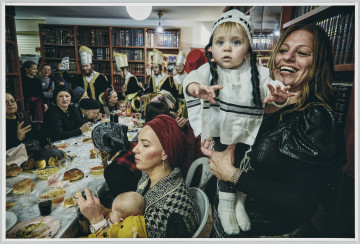 scena rodzajowa - Poziomy, barwny kadr fotograficzny przedstawiający ucztującą grupę kobiet, mężczyzn i dzieci we wnętrzu. Na pierwszym planie, po prawej stronie, widać dwie kobiety z niemowlętami – jedna z nich siedzi przy stole trzymając dziecko na kolanach, druga stoi, śmiejąc się prezentuje na rękach niemowlę w kostiumie przebierańców. Jej sylwetka zajmuje całą prawą krawędź kadru. Od lewego narożnika, pod kątem, w głąb sceny biegnie długi stół zastawiony jedzeniem i piciem. Po obu jego stronach siedzą kobiety i dzieci, za nim widać postaci 4 mężczyzn o fryzurach z charakterystycznymi długimi, zakręconymi pasmami nad uszami (pejsami) poprzebieranych w jednakowe kostiumy. W głębi, po lewej stronie stoi kilka kobiet.