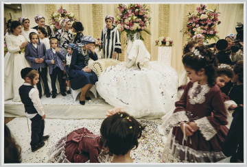 scena rodzajowa - Poziomy, barwny kadr fotograficzny przedstawiający scenę ślubną. W środku kadru na jasnym szezlongu siedzi panna młoda w białej, sukni ślubnej o szerokiej spódnicy. Kobieta ma twarz zasłoniętą szczelnie nieprzejrzystą białą chustą/welonem. Po jej prawej stronie siedzi kobieta eleganckiej granatowej sukience, ze sznurem pereł na szyi i kwiecistej chuście na głowie. Wspiera głowę na lewej ręce, dłonią zasłania twarz w geście zmartwienia/rozpaczy. Za nią stoi grupka kobiet w odświętnych sukienkach, z chustami na głowach oraz małych chłopców w podobnych garniturach, z charakterystycznymi fryzurami (krótkie włosy oraz długie skręcone pasma włosów zakrywające uszy – pejsy) oraz płaskich, okrągłych nakryciach głowy (jarmułki). Cała ta grupa znajduje się na podeście, na tle ozdobnej, jasnej zasłony oraz dużych bukietów kolorowych kwiatów. Poniżej podestu znajduje się grupa dzieci – chłopiec i kilka dziewczynek w odświętnych strojach (dziewczynki w takich samych sukienkach). Za nimi, w prawym, górnym rogu kadru widać kilka kobiet w chustach na głowach oraz aparat fotograficzny z dużym obiektywem zasłaniający trzymającego go fotografa. Suknia panny młodej, podest i podłoga zasypane są kolorowy konfetti.