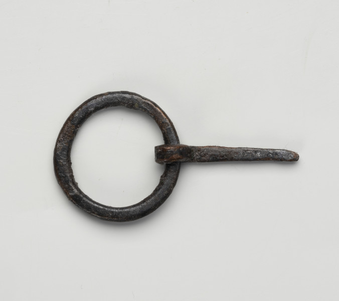 sprzączka do pasa - Ujęcie z góry; Kolista sprzączka żelazna ze spłaszczonego drutu z ruchomym kolcem na ramie. Kolec przymocowano poprzez wygięcie tępego końca drutu i owiniecie go wokół obręczy sprzączki. Na powierzchni zachowane są pojedyncze włókna.