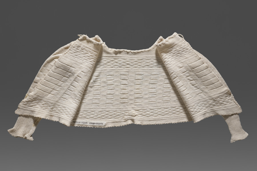 Kaftanik dziecięcy w kolorze écru. Ścieg mieszany, układającymi się w poziome pasy z dominującym motywem szachownicy. W centralnej części wzór jest wyraźnie większy, Wzdół dolnej krawędzi i na wysokości piersi drobniejszy. Poły kaftanika są wydłużone. Kaftanik wiązany pod szyją na taśmę w tym samym kolorze. Rękawy zakończone ściągaczem.