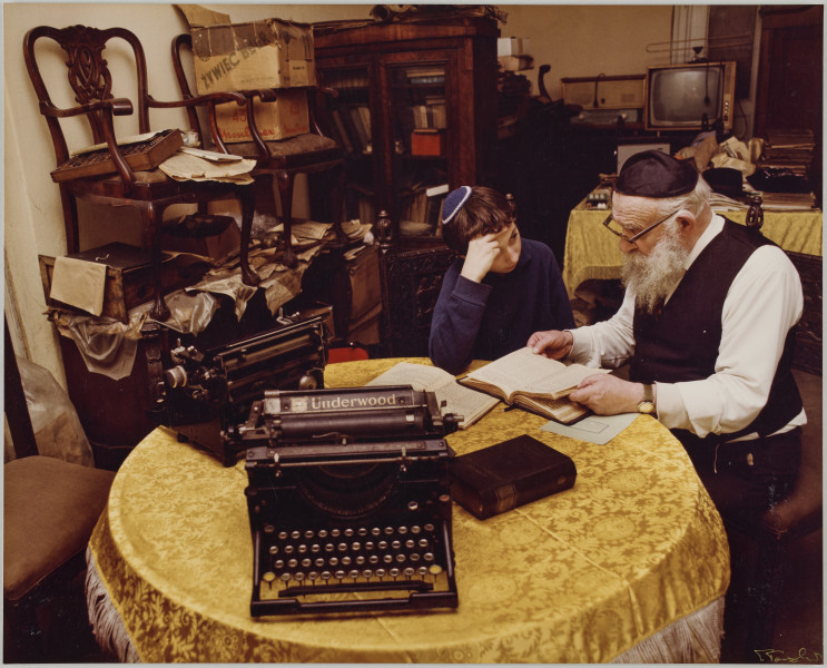 fotografia - Zdjęcie przedstawia starszego mężczyznę z siwą brodą i kilkunastoletniego chłopca siedzących przy okrągłym, nakrytym żółtym obrusem stole. Mężczyzna ma na sobie białą koszulę, czarną kamizelkę, jarmułkę i okulary. Chłopiec ma na sobie ciemny sweter i jarmułkę. Starszy mężczyzna siedzi po prawej stronie kadru. Przed nim leżą książki. Jedną z nich trzyma otwartą w dłoniach. Jest zwrócony twarzą do chłopca. Chłopiec (siedzący po lewej stronie) podpiera czoło ręką, którą opiera łokciem o stół. Patrzy na otwartą książkę. Oprócz książek, na stole znajdują się także dwie maszyny do pisania. W tle widać meble stanowiące wyposażenie pokoju. Na części z są rozłożone gazety lub stoją kartony. 