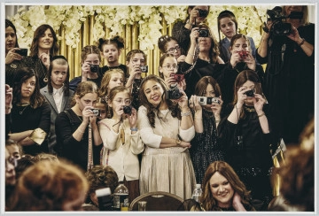 scena rodzajowa - Poziomy, barwny kadr fotograficzny przedstawiający scenę we wnętrzu. Na tle ściany ozdobionej jasna, dekoracyjną kotarą i girlandami jasnych, sztucznych kwiatów stoi grupa kobiet i dziewcząt oraz dwóch chłopców. Wszyscy są ubrani z odświętne stroje, chłopcy mają charakterystyczne fryzury – krótkie włosy i długie, spiralne pasma opadające na uszy (pejsy). Wszyscy patrzą w jednym kierunku, przed siebie, część z dziewcząt trzyma aparaty fotograficzne w geście fotografowania.
