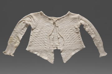 Kaftanik dziecięcy w kolorze écru. Ścieg mieszany, układającymi się w poziome pasy z dominującym motywem szachownicy. W centralnej części wzór jest wyraźnie większy, Wzdół dolnej krawędzi i na wysokości piersi drobniejszy. Poły kaftanika są wydłużone. Kaftanik wiązany pod szyją na taśmę w tym samym kolorze. Rękawy zakończone ściągaczem.