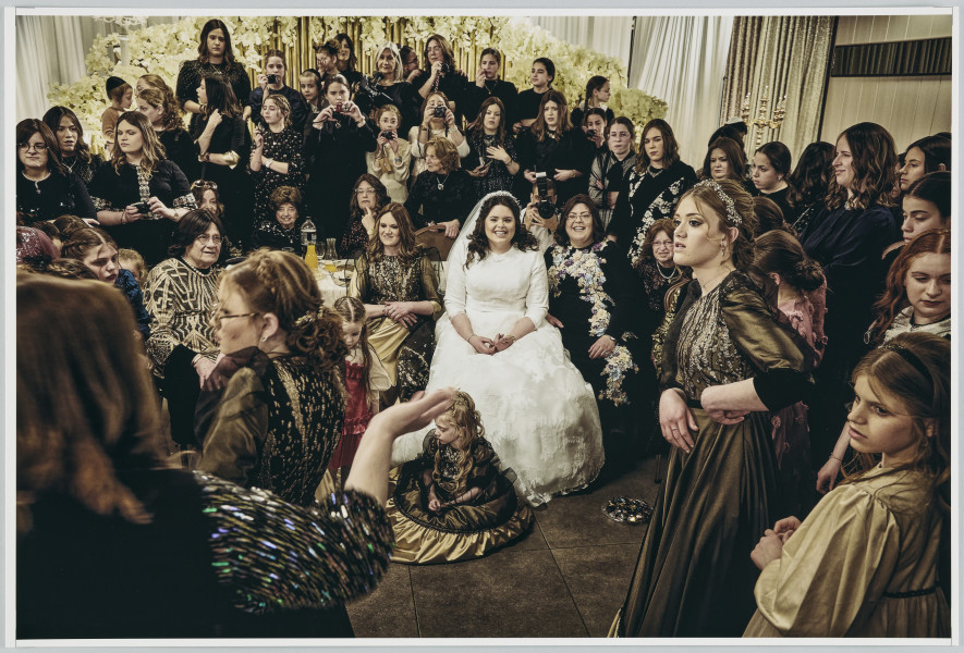 scena rodzajowa - Poziomy, barwny kadr fotograficzny przedstawiający pozowaną scenę ślubną z udziałem dużej grupy kobiet. Na pierwszym planie, po lewej stronie, tyłem do widza stoi kobieta - widoczny jest tylko fragment jej sylwetki. Prawą rękę trzyma wzniesioną gestykulując. W centralnej części kadru widać we fragmencie stół nakryty białym obrusem. Przed nim, po środku siedzi uśmiechnięta panna młoda, obok niej inne kobiety. Za stołem siedzą starsze kobiety. Z tyłu i dookoła stołu stoją pozostałe uczestniczki sceny. Wszystkie są zwrócone w stronę kobiety z pierwszego planu. Część z nich ma aparaty fotograficzne. Pośród zebranych jest także jeden mężczyzna robiący zdjęcie.