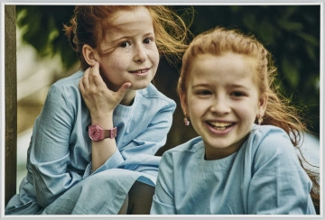 scena rodzajowa - Poziomy, barwny kadr fotograficzny przedstawiający dwie uśmiechnięte dziewczynki o rudych, długich włosach rozwiewanych przez wiatr. Dzieci siedzą blisko siebie, jedna na pierwszym planie, druga tuż za nią. Obie są ubrane w jednakowe niebieskie sukienki. Dziewczynka z tyłu ma na lewej ręce różowy zegarek.