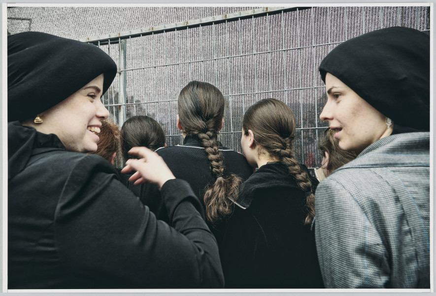 scena rodzajowa - Poziomy, barwny kadr fotograficzny przestawiający dwie zwracające się do siebie kobiety (rozmawiające?), ubrane w okrycia wierzchnie w stonowanych kolorach oraz czarne nakrycia głowy. Kobieta stojąca po lewej unosi prawą rękę. Przed nimi widać tłoczące się dziewczynki w ciemnych ubraniach i z włosami splecionymi w warkocze, stojące tyłem do widza. Grupa patrzy w kierunku kraty i wiszącej przed nią półprzezroczystej zasłony.