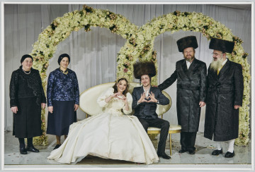 scena rodzajowa - Poziomy, barwny kadr fotograficzny, na którym w centralnej części widoczna jest para młoda siedząca na ozdobnej ławce. Panna młoda ubrana jest białą ozdobną suknię, złoty wianek, zaś mężczyzna w biała koszulę, ciemna marynarkę, spodnie i buty. Na głowie ma dużą czapkę formie walca wykonaną z futra. Każdy z nowożeńców palcami pokazuje gest serca. Po lewej stronie stoją dwie kobiety ubrane w odświętne garsonki oraz naszyjniki oraz czarne chustki na głowach. Po prawej stronie – stoi 2 mężczyzn ubranych w białe koszule, długie połyskliwe, odświętne płaszcze (chałaty), oraz podobne, jak pan młody czapki. Wszyscy znajdują się na tle dużej stojącej ślubnej dekoracji z girland kwiatowych i białej zasłony.