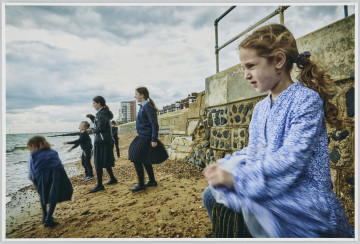 scena rodzajowa - Poziomy, barwny kadr fotograficzny przedstawiający sylwetki dzieci bawiących się nad morzem przy pochmurnej pogodzie. Na pierwszym planie siedzi mała dziewczynka z rudymi, długimi włosami spiętymi z tyłu głowy, ubrana w niebieską sukienkę w białe drobne kwiatuszki i szare rajstopy. Na planie dalszym czwórka dzieci stojących i patrzących w kierunku morza. Jedna dziewczyna trzyma uniesioną rękę, w której coś trzyma (kamień?). Za nimi widać sylwetkę chłopca odwróconego do grupy tyłem. W tle mieszkalne bloki o różnej wysokości. Ostrość nastawiona została na twarz dziewczynki na pierwszym planie, dlatego inne elementy zdjęcia są rozmyte.